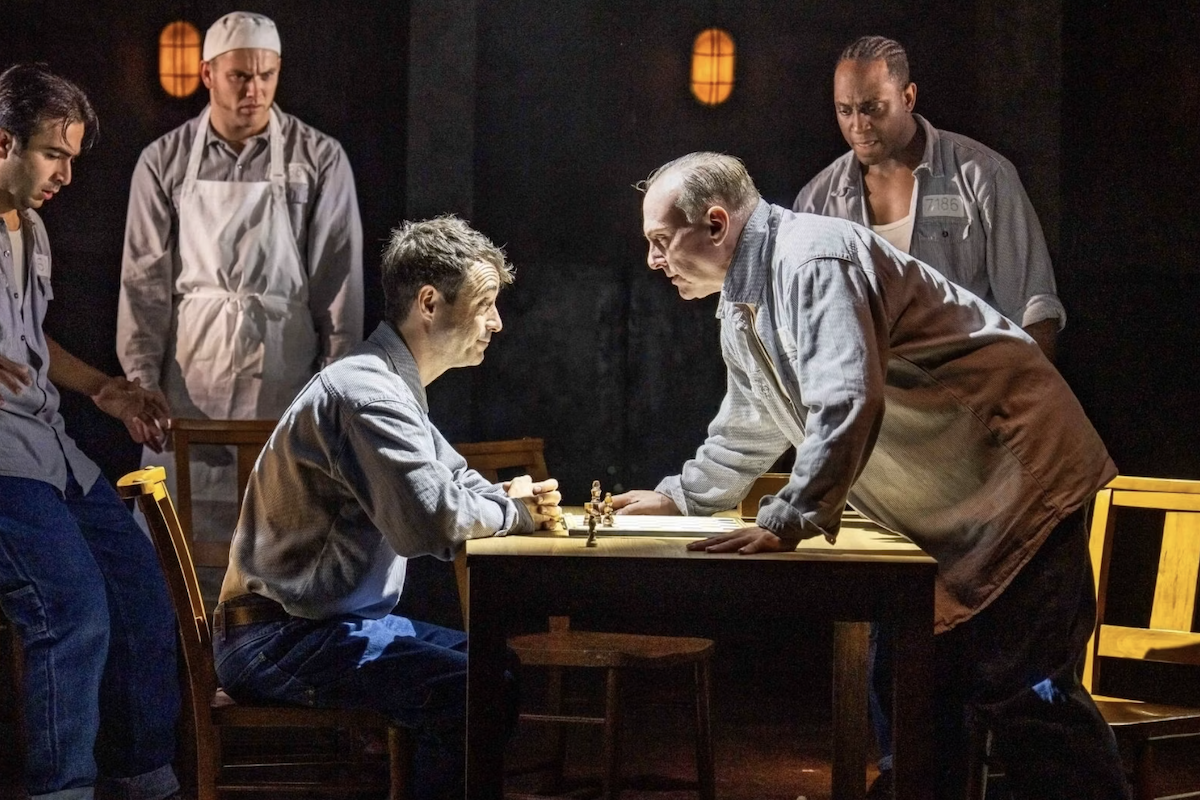 Actors play chess in prison uniforms during a stage production of The Shawshank Redemption as other inmates watch intensely in dim light.